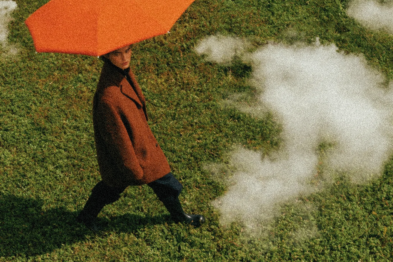 Person in a brown coat walking across grass under an orange umbrella, with film grain across the frame