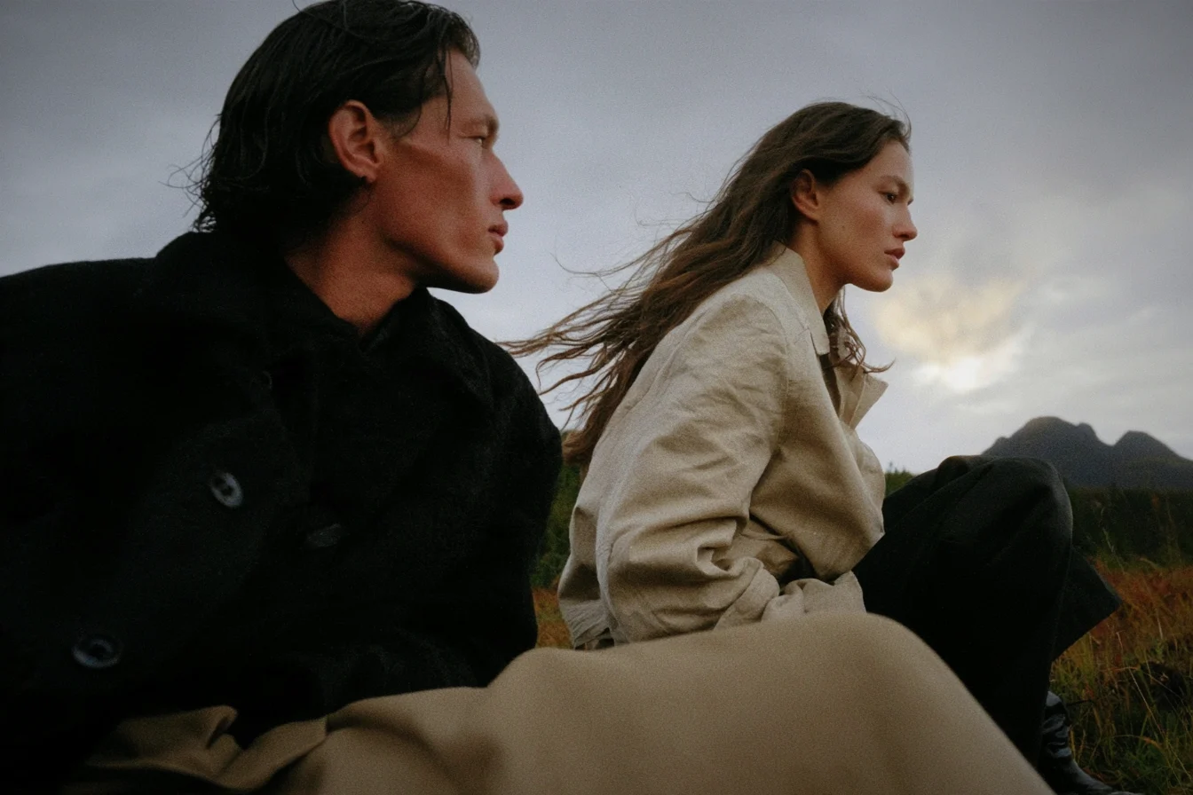 Two people in dark and beige coats on a hillside under a cloudy sky, with a vignette darkening the edges